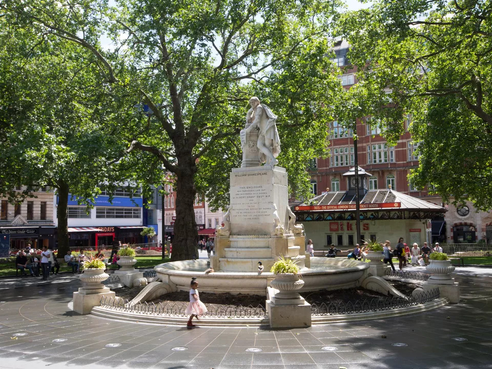 A decorative fountain stands in the center of a bustling city square, surrounded by people and buildings